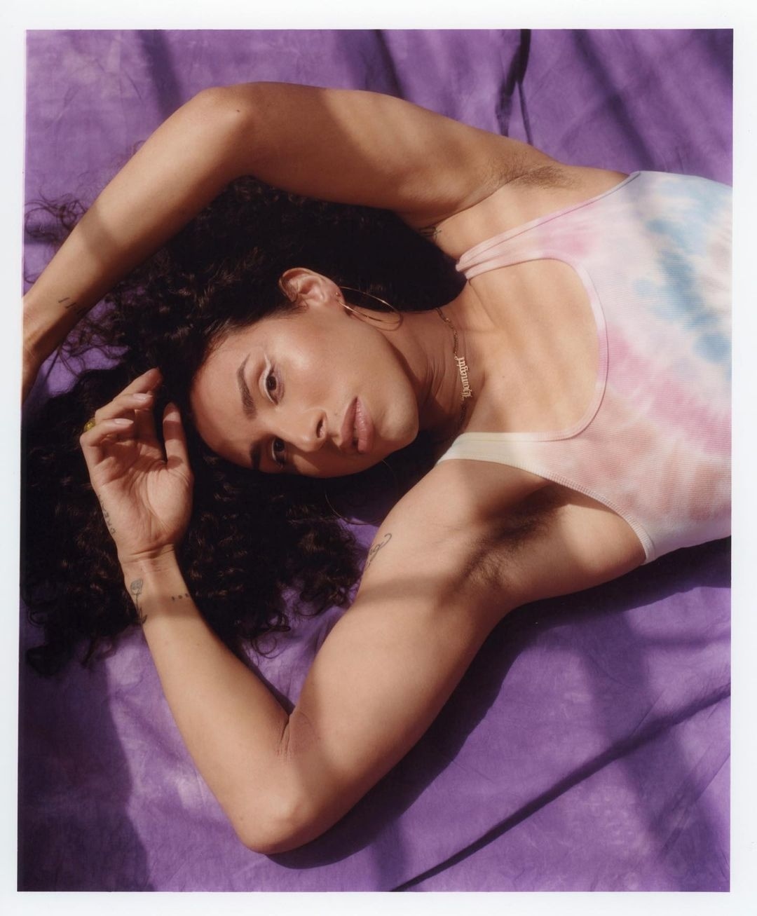 Attractive adult laying on a purple surface, facing the camera. A tie-dye tank top and gold necklace complete the look, accented by shadows.