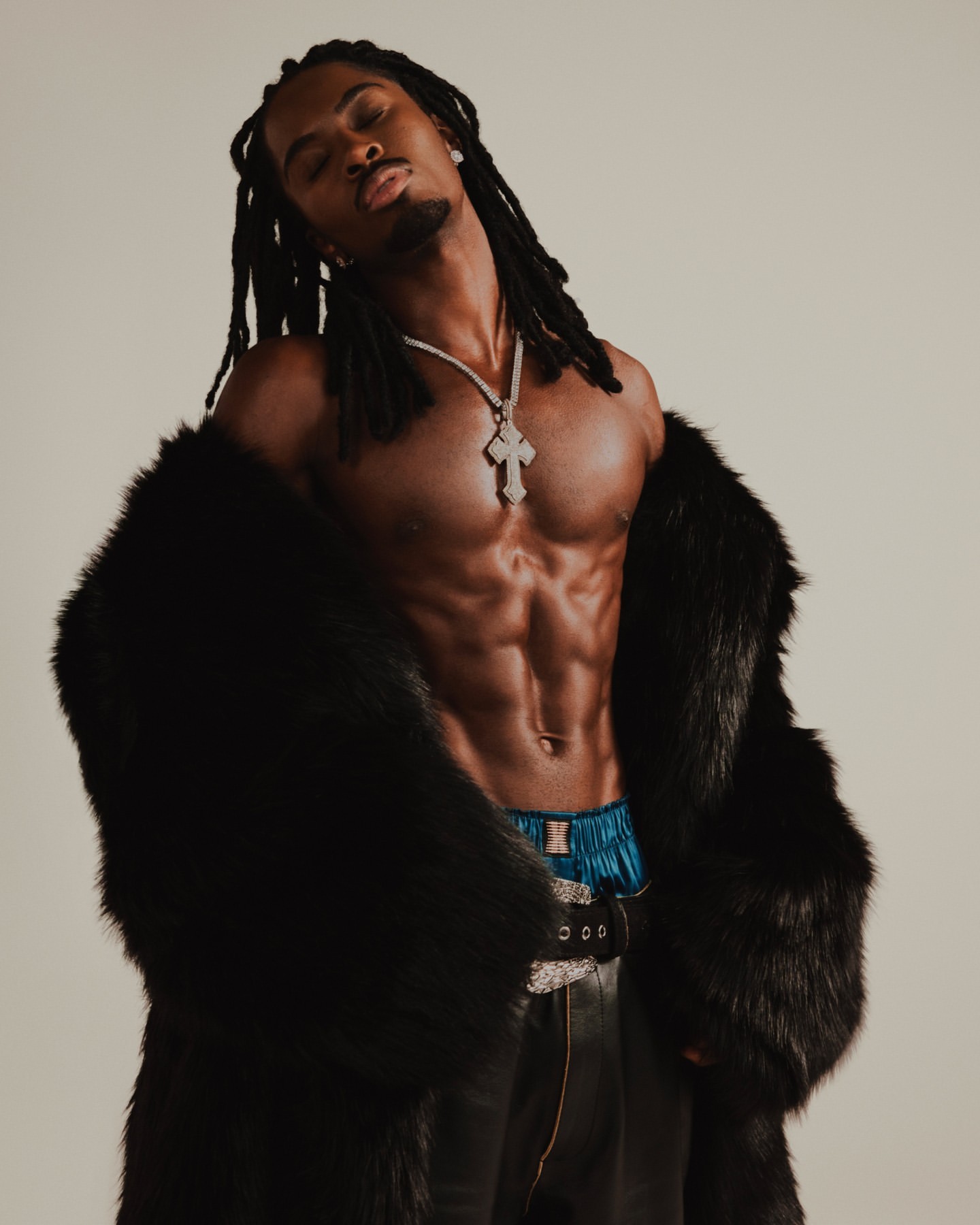 Muscular Black man with dreadlocks poses shirtless wearing a fur coat, necklace, and leather pants against a neutral studio background.
