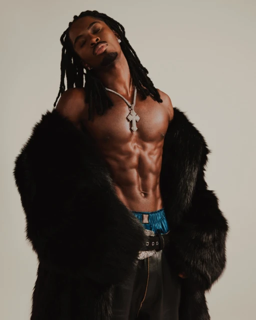 Muscular Black man with dreadlocks poses shirtless wearing a fur coat, necklace, and leather pants against a neutral studio background.