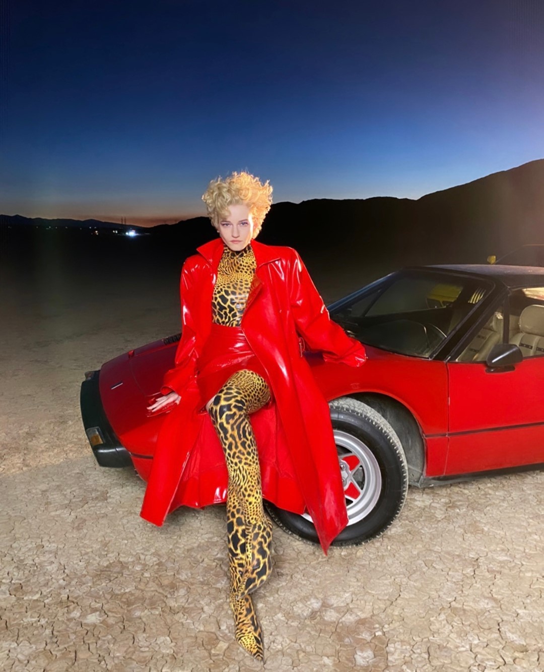 Woman in a red coat and leopard print tights leans on a red sports car in a desert landscape against a dusk sky.
