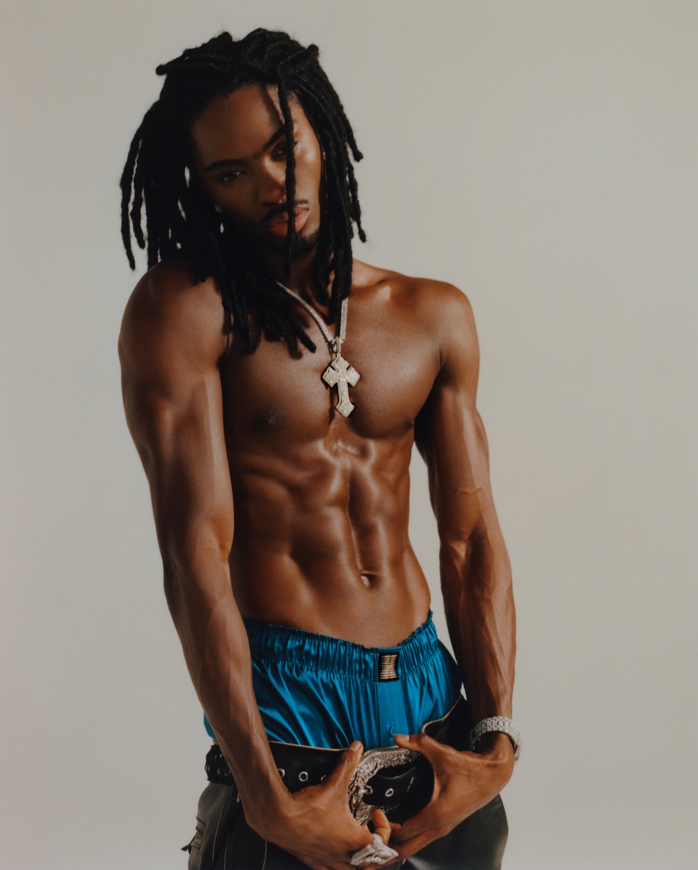 Shirtless man with dreadlocks poses in front of a solid background, wearing a cross necklace, shorts, and a belt.