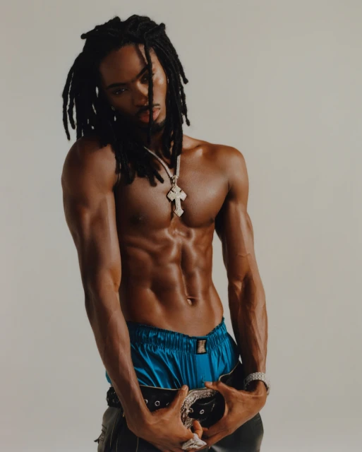 Shirtless man with dreadlocks poses in front of a solid background, wearing a cross necklace, shorts, and a belt.
