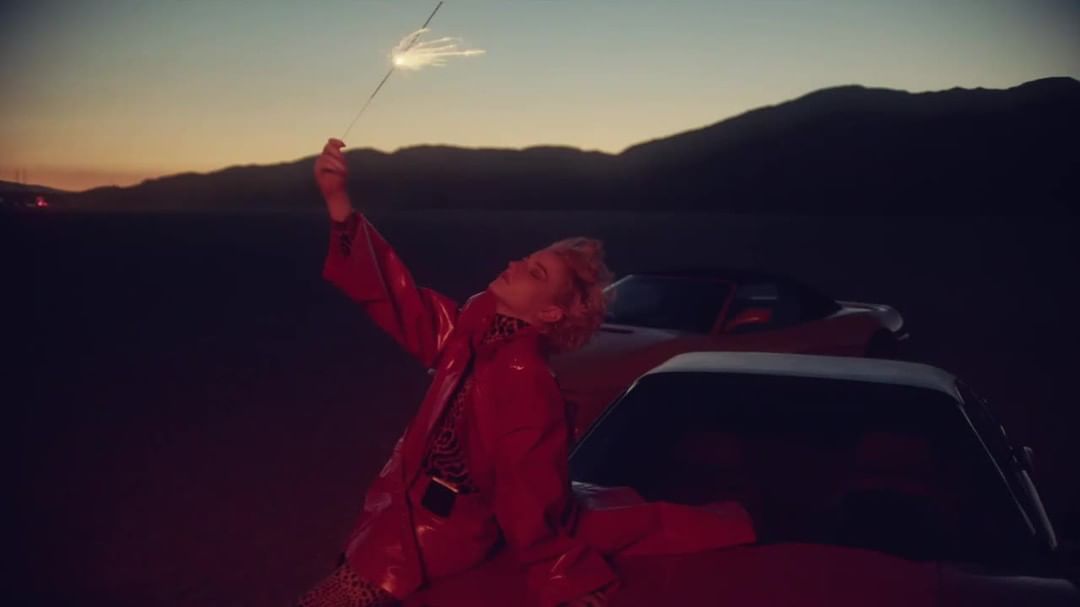 Young woman in red jacket holding sparkler at sunset in desert near vintage cars. Mountains in the background. Golden hour lighting.
