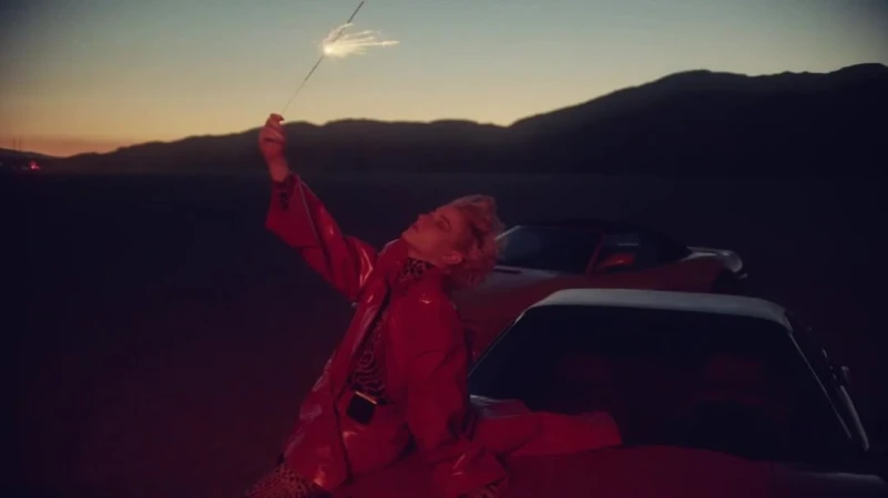 Young woman in red jacket holding sparkler at sunset in desert near vintage cars. Mountains in the background. Golden hour lighting.