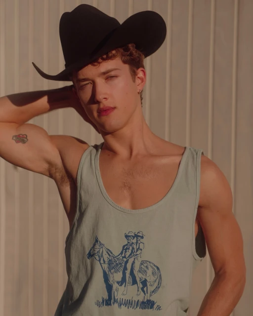 Portrait of a young adult male in a cowboy hat and graphic tank top against a neutral wall, in soft natural light.