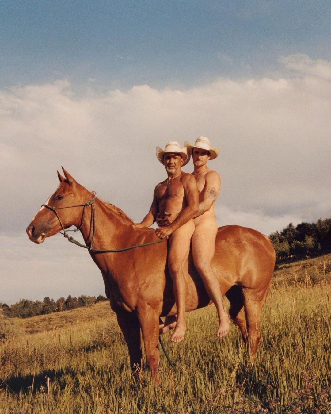 Two nude adult males wearing cowboy hats riding a chestnut horse through a grassy field under a blue sky with scattered clouds.