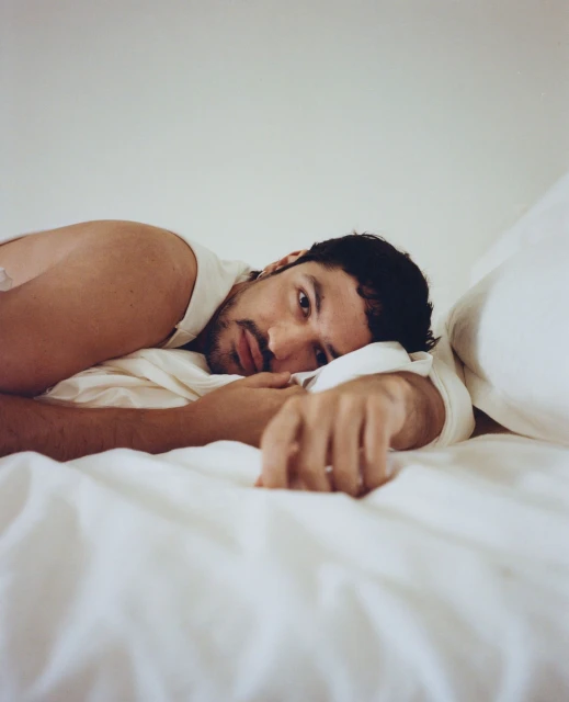 A man with dark hair and a slight beard reclines on a white bed, looking towards the camera with a soft expression. The lighting is soft.