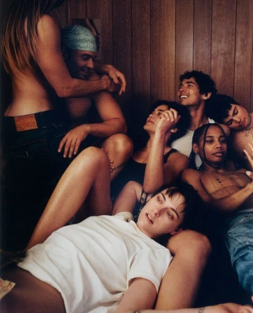 Seven young adults gathered indoors, some embracing and reclining, with a background of wood panels and soft lighting in an intimate setting.