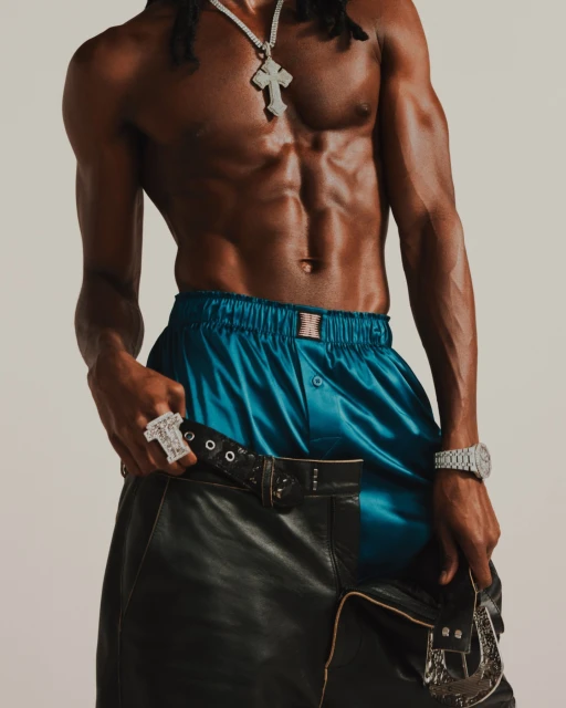 Athletic Black male model with jewelry poses shirtless in blue shorts and leather chaps against a neutral backdrop in a studio shot.