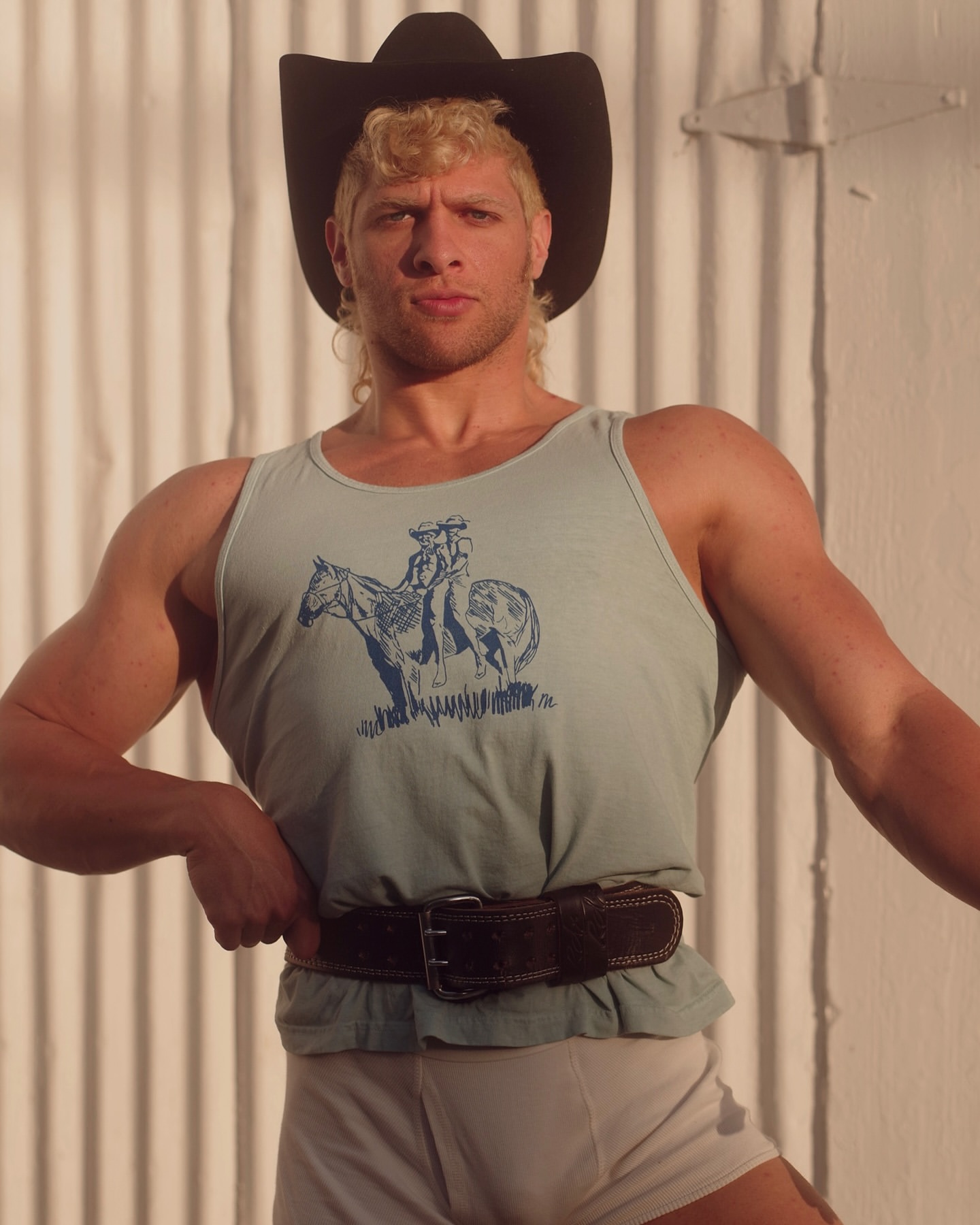 Muscular man in cowboy hat and tank top with horse graphic poses indoors against corrugated white wall, lit by natural light.