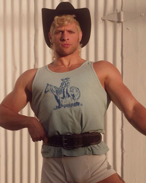 Muscular man in cowboy hat and tank top with horse graphic poses indoors against corrugated white wall, lit by natural light.