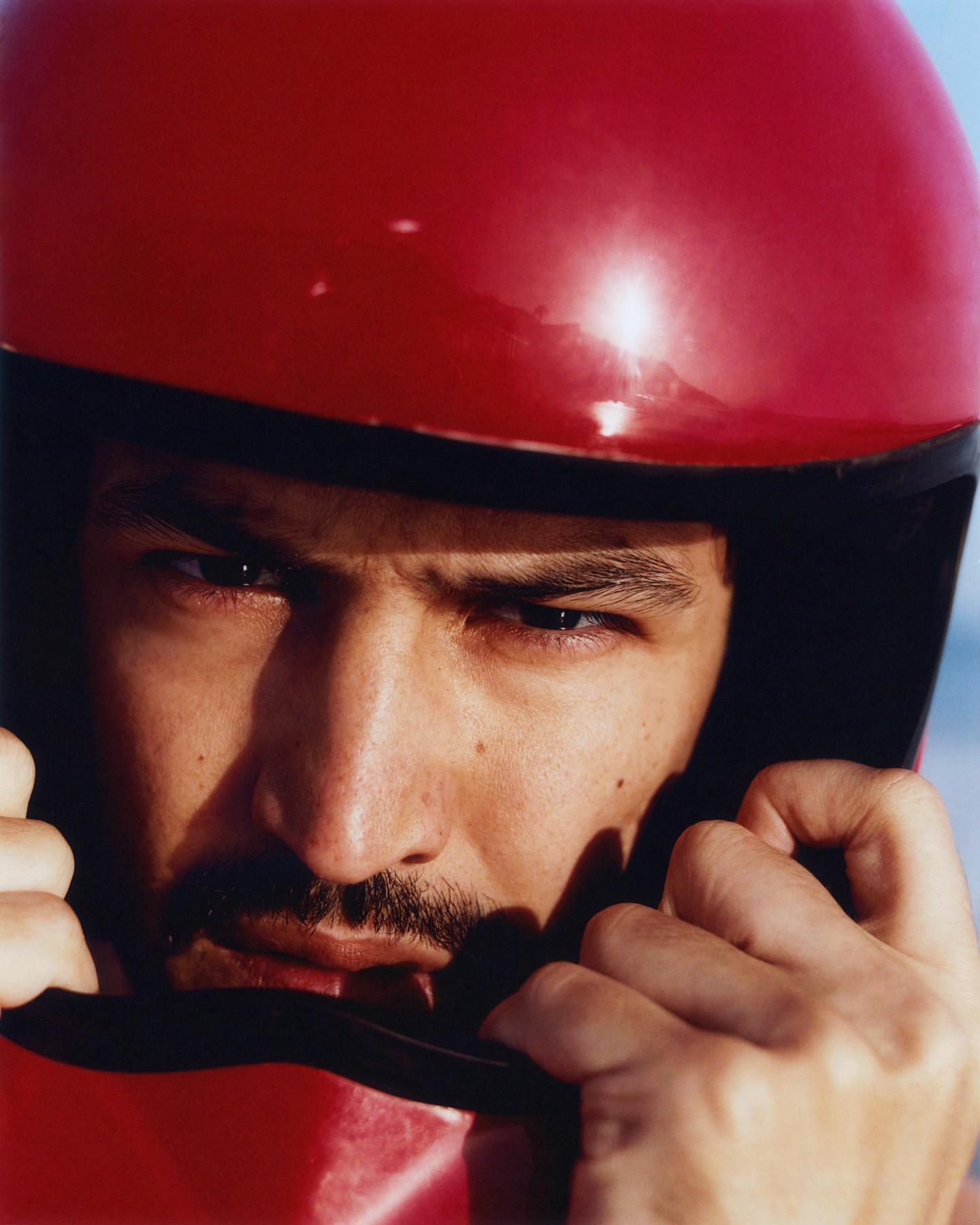 Close-up of a man wearing a red full-face helmet, gazing intently at the viewer with a determined expression, his hands near the helmet.