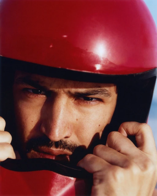 Close-up of a man wearing a red full-face helmet, gazing intently at the viewer with a determined expression, his hands near the helmet.