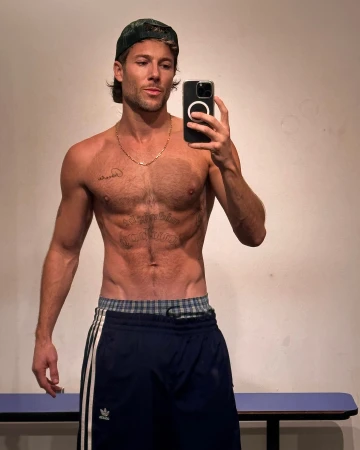 Athletic man with tattoos takes a selfie in gym shorts and a baseball cap, showing off his physique in an indoor setting.