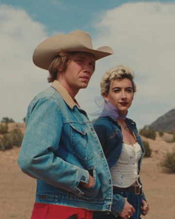 Man in cowboy hat and denim jacket standing outdoors beside a woman with short blonde hair against a desert landscape.