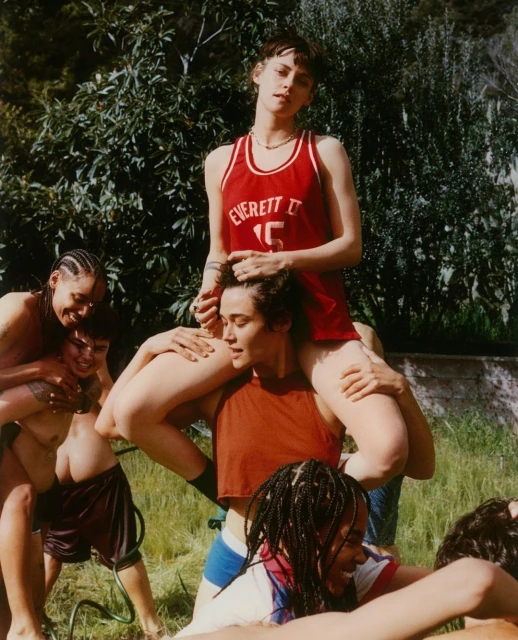Group of young adults outdoors; one woman is being carried on another woman's shoulders, others are behind with trees in the background.