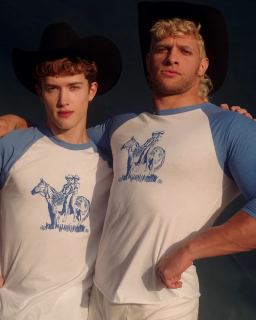 Two adult males wear matching cowboy-themed shirts and black hats in a portrait, posing together against a dark background.