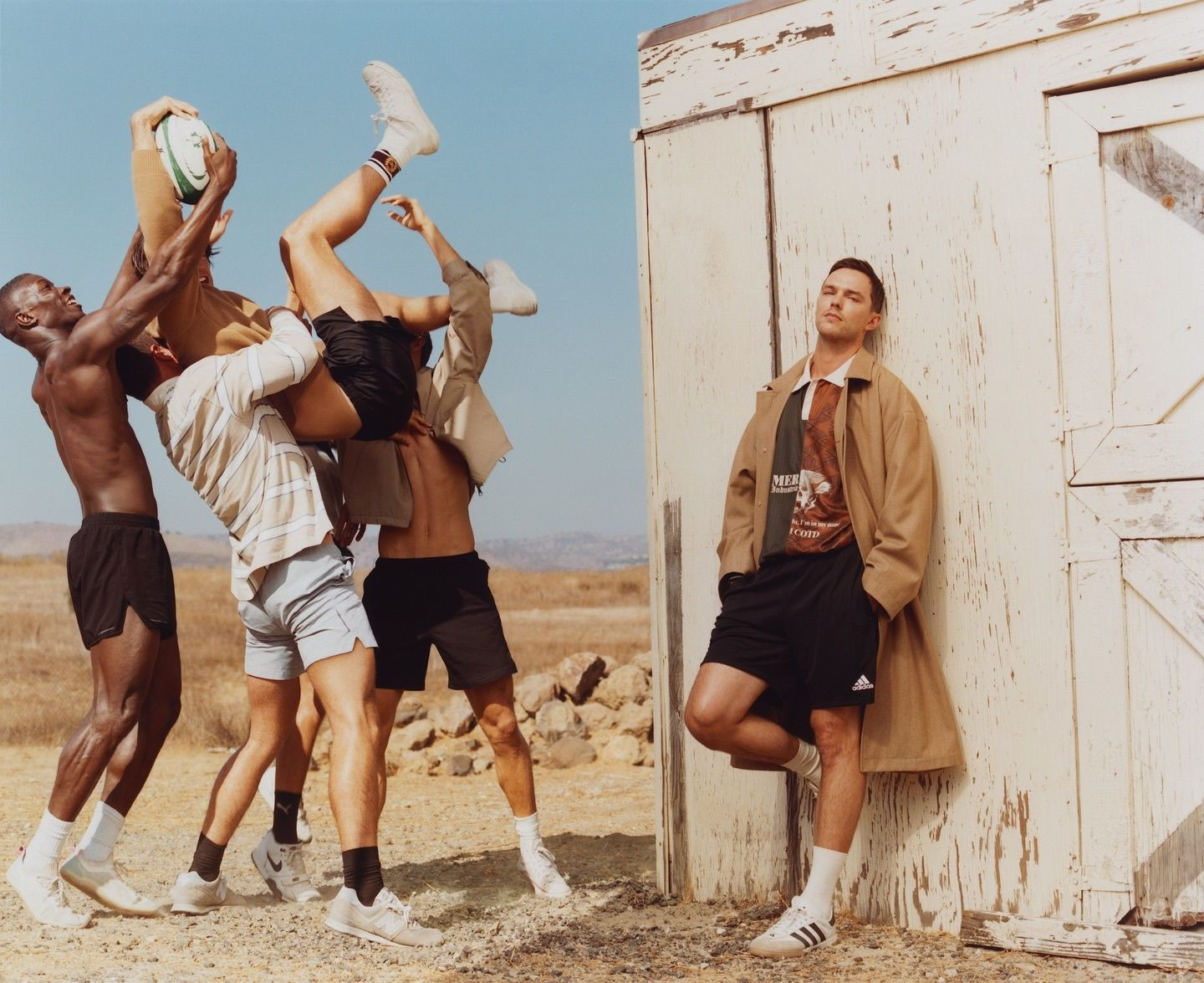 A group of athletic men play rugby outdoors under a clear sky, one man standing apart in a beige coat, Adidas shorts, and white sneakers.