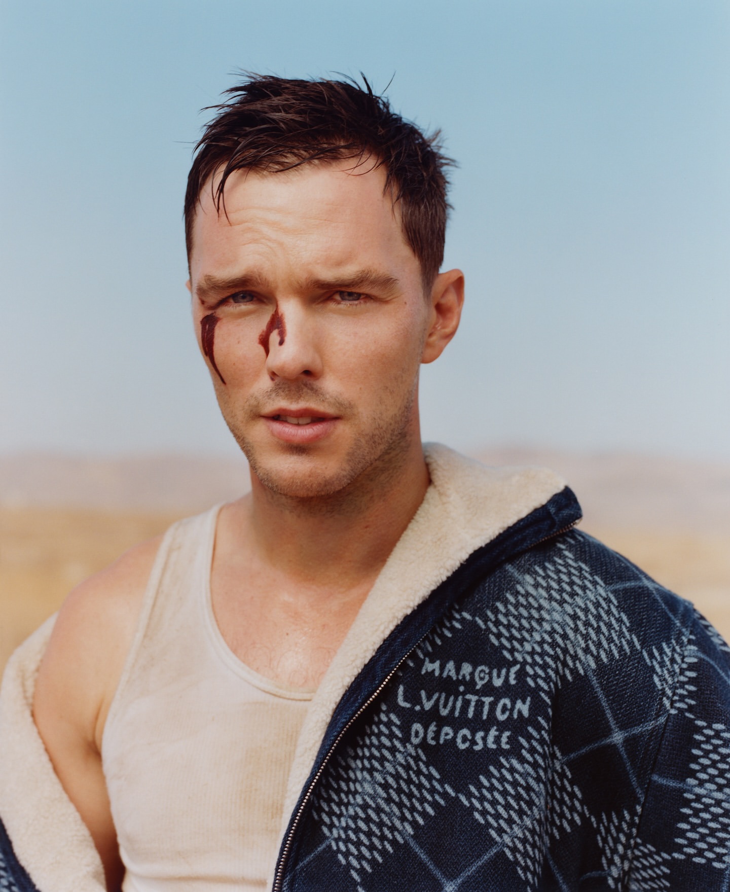 Man with simulated blood on his face, wearing a tank top and denim jacket, posing against a desert and sky backdrop for a portrait.