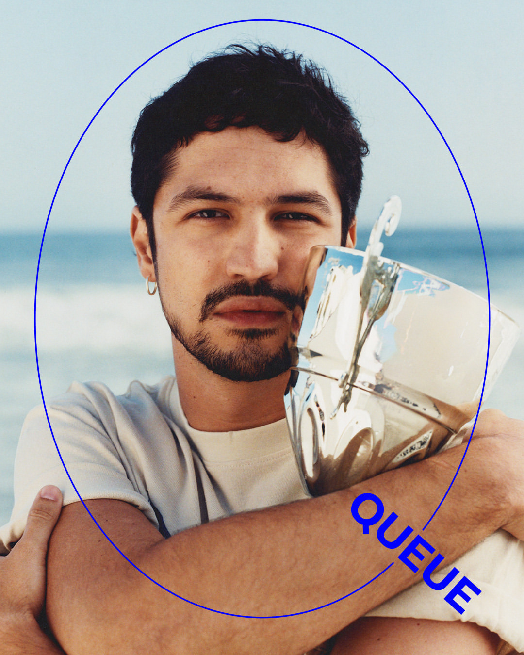 Portrait of a man holding a reflective silver trophy against a beach background, wearing a light t-shirt and silver hoop earrings.