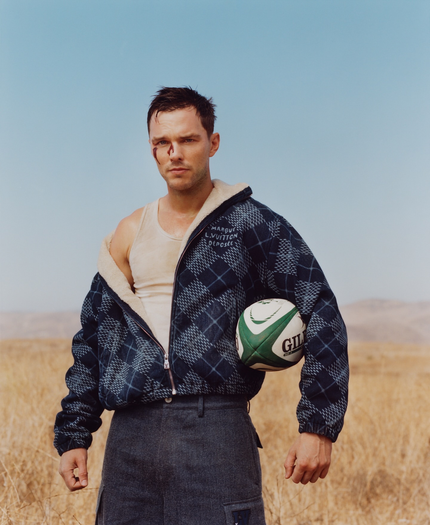 Male model with facial cuts poses outdoors wearing a blue plaid jacket, white tank top, and grey pants while holding a rugby ball.
