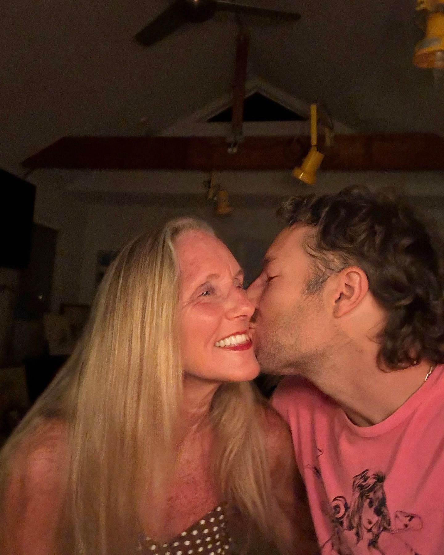 Close-up of a smiling mature woman being kissed on the cheek by a younger adult male indoors, under dim lighting.