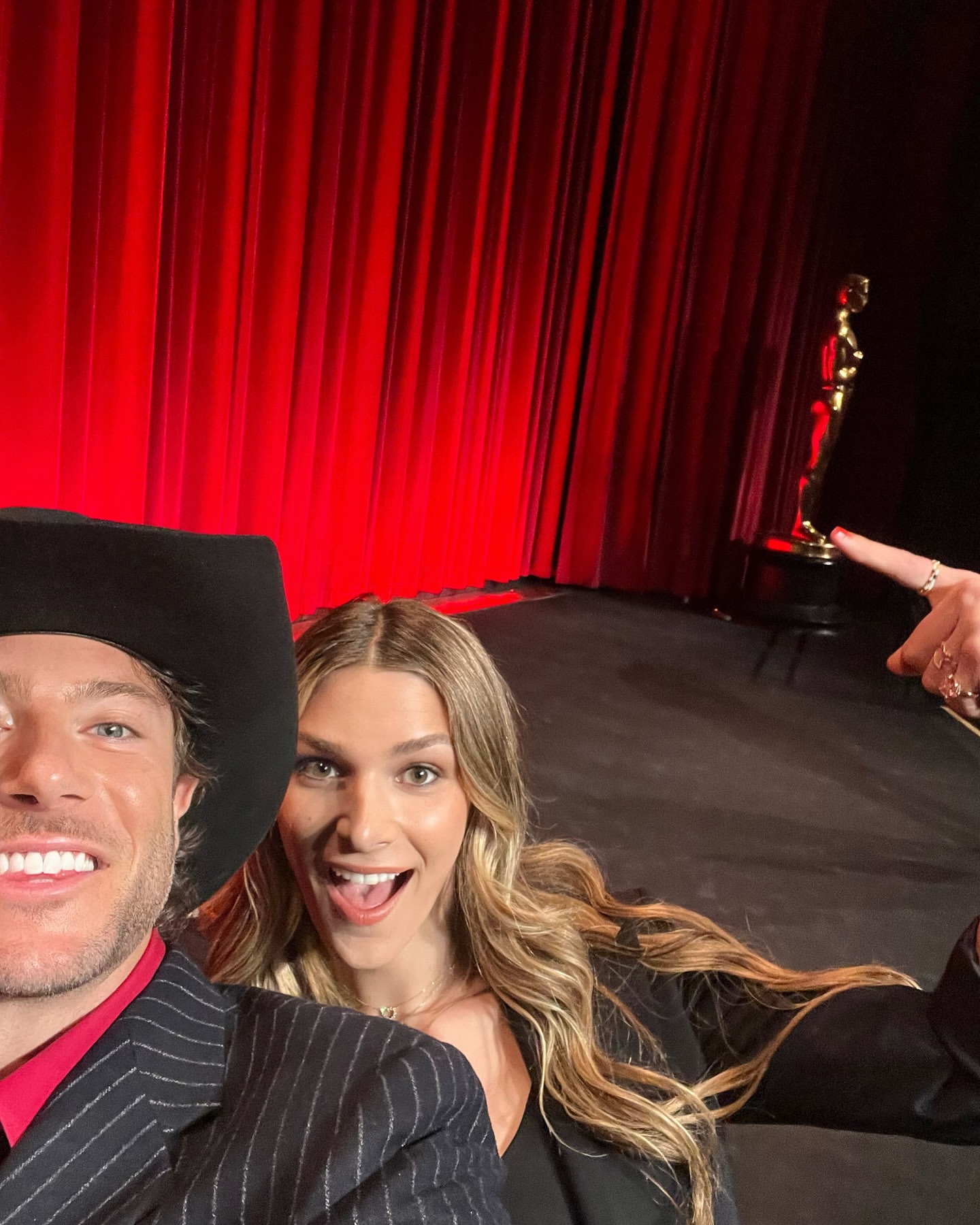 Two people pose for a selfie on a stage in front of red curtains, with an award statue visible in the background. Smiling, formal wear.