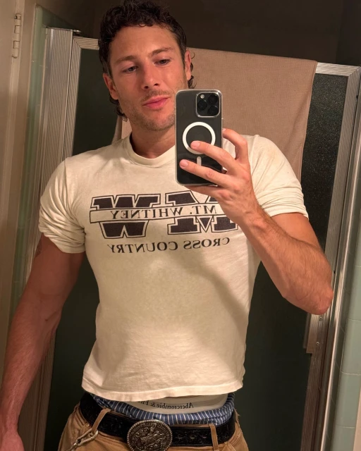 Indoor selfie of adult male in a bathroom wearing a Mt. Whitney t-shirt, tan pants, and decorative belt, holding a smartphone.
