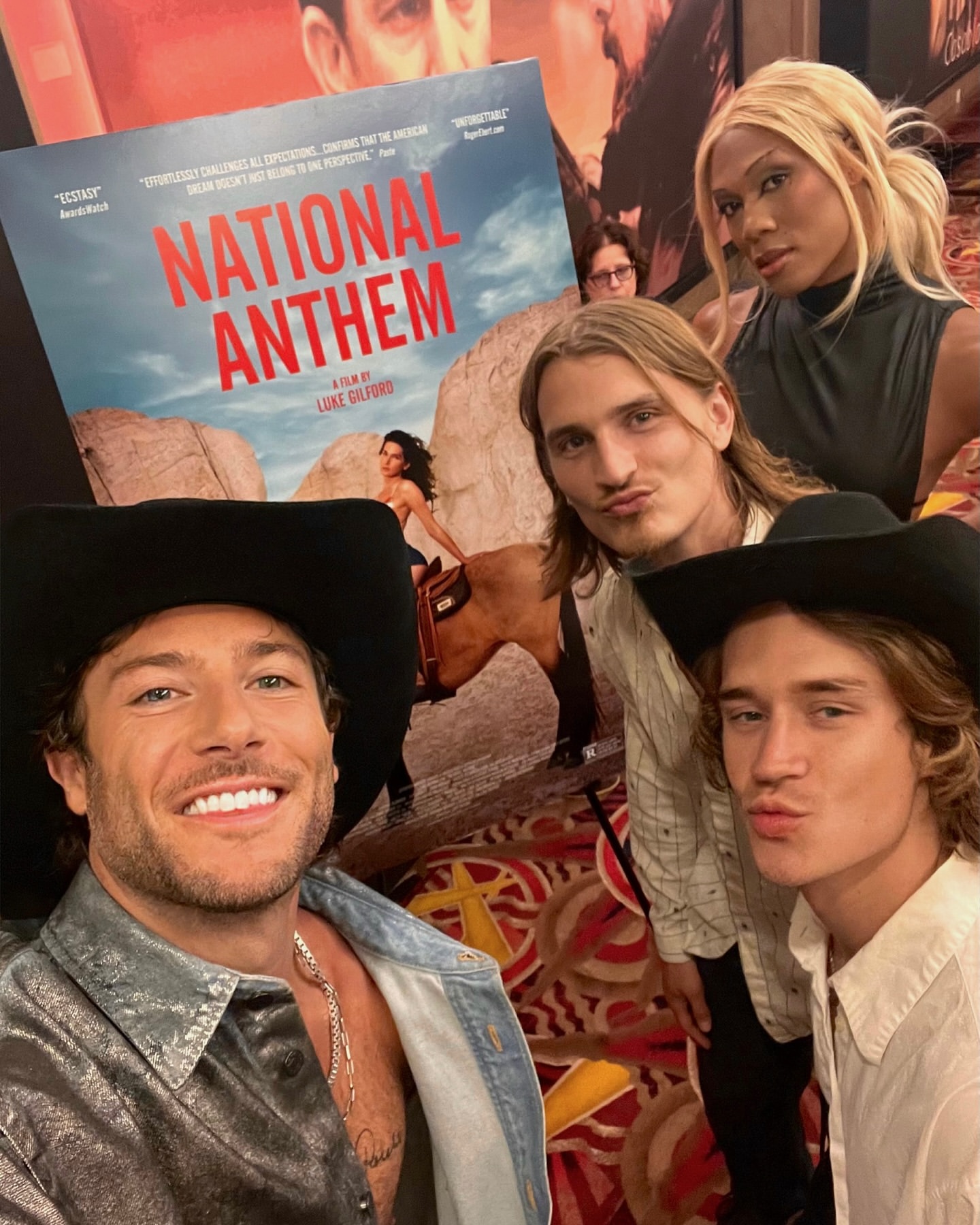 Group selfie with four young adults in front of a movie poster for "National Anthem," featuring a woman on horseback.