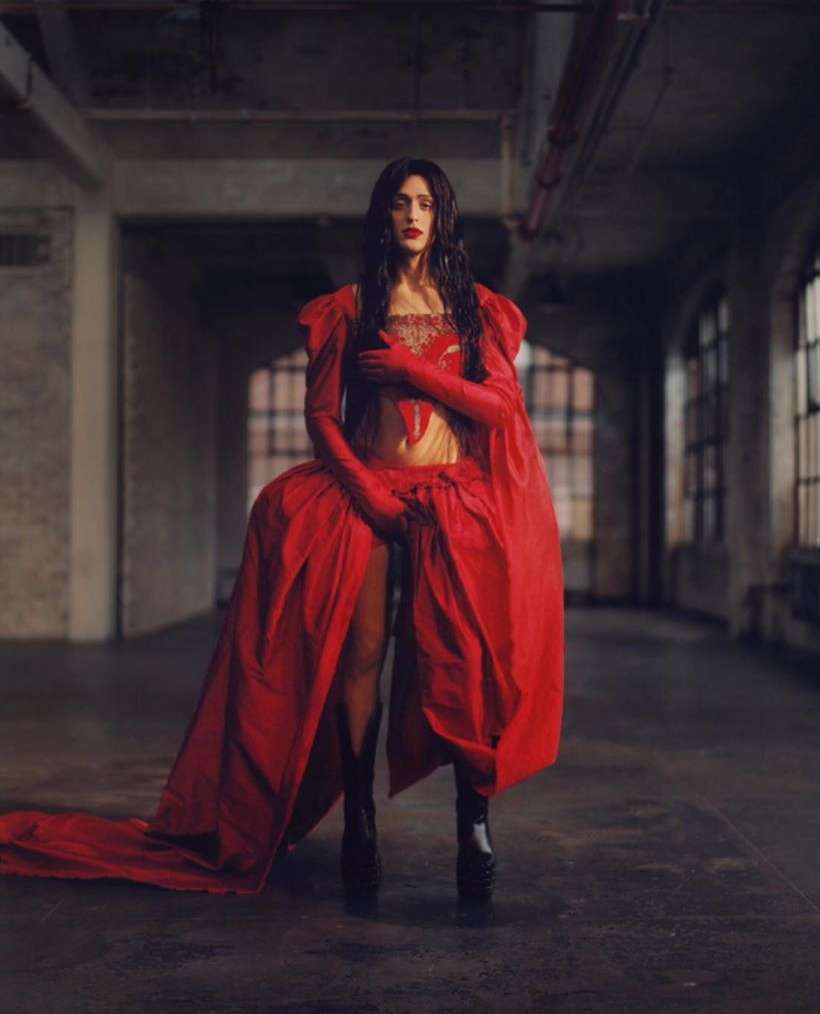 A woman poses indoors in a vibrant red gown with long sleeves and black platform boots against a grey brick industrial backdrop.