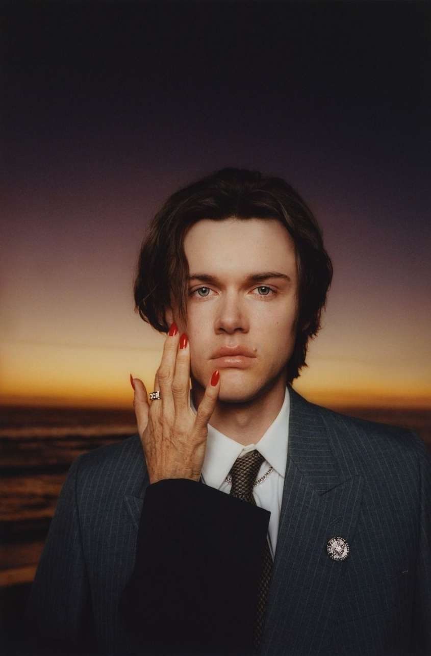 Portrait of a fair-skinned young adult with brown hair wearing a pinstripe suit, with an older hand with red nails on their face, set on a beach.
