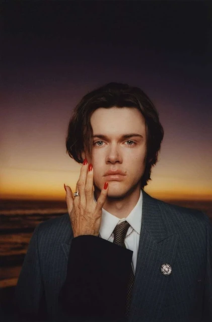 Portrait of a fair-skinned young adult with brown hair wearing a pinstripe suit, with an older hand with red nails on their face, set on a beach.