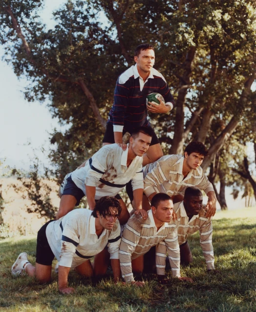 A human pyramid of six rugby players outdoors on green grass under trees. One player stands holding a ball on top of the pyramid.
