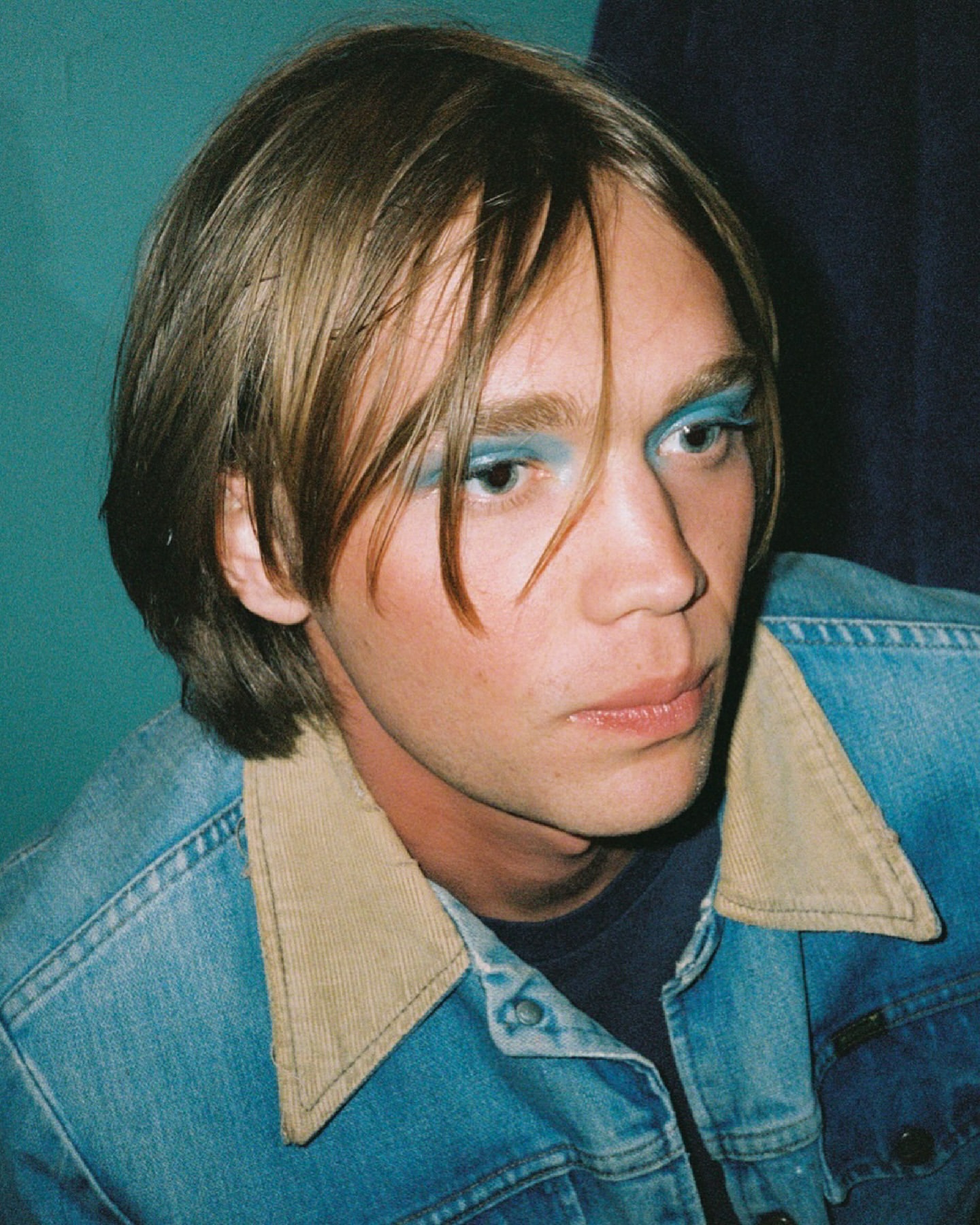 Portrait of a man wearing blue eyeshadow and a denim jacket with a tan corduroy collar against a teal wall.
