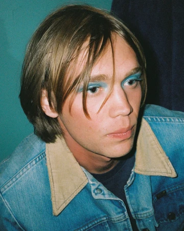 Portrait of a man wearing blue eyeshadow and a denim jacket with a tan corduroy collar against a teal wall.