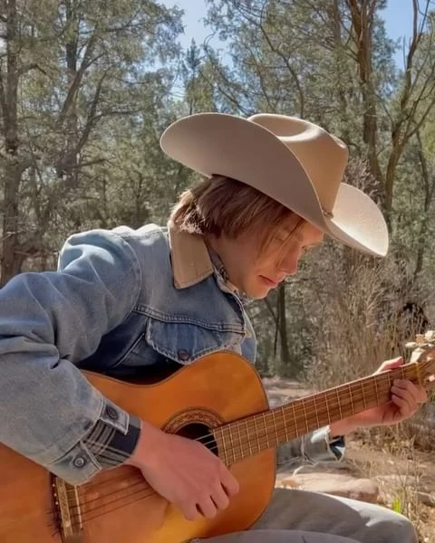 Young adult wearing a cowboy hat playing an acoustic guitar in an outdoor setting with trees in the background.