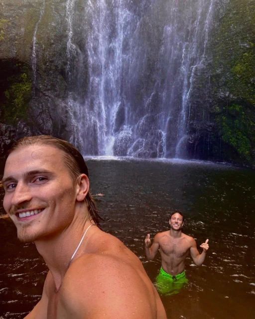 Two adult men are standing in the water in front of a cascading waterfall. They are shirtless and smiling in the outdoor scene.