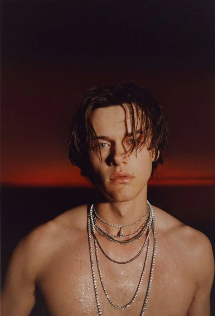 Striking portrait of a young man with dark hair wearing layered necklaces against a diffused red and dark background, chest-up shot.