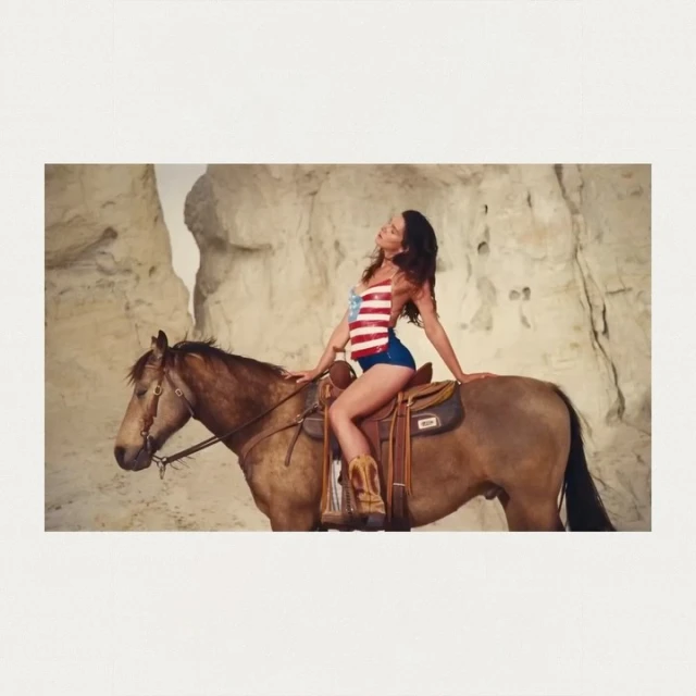 A woman with long dark hair rides a brown horse in a desert setting wearing a USA flag top and denim shorts. Neutral color palette.