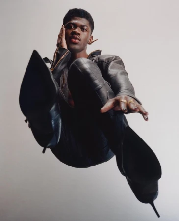 Low angle portrait of Lil Nas X wearing a dark leather jacket and high heeled boots against a white backdrop.