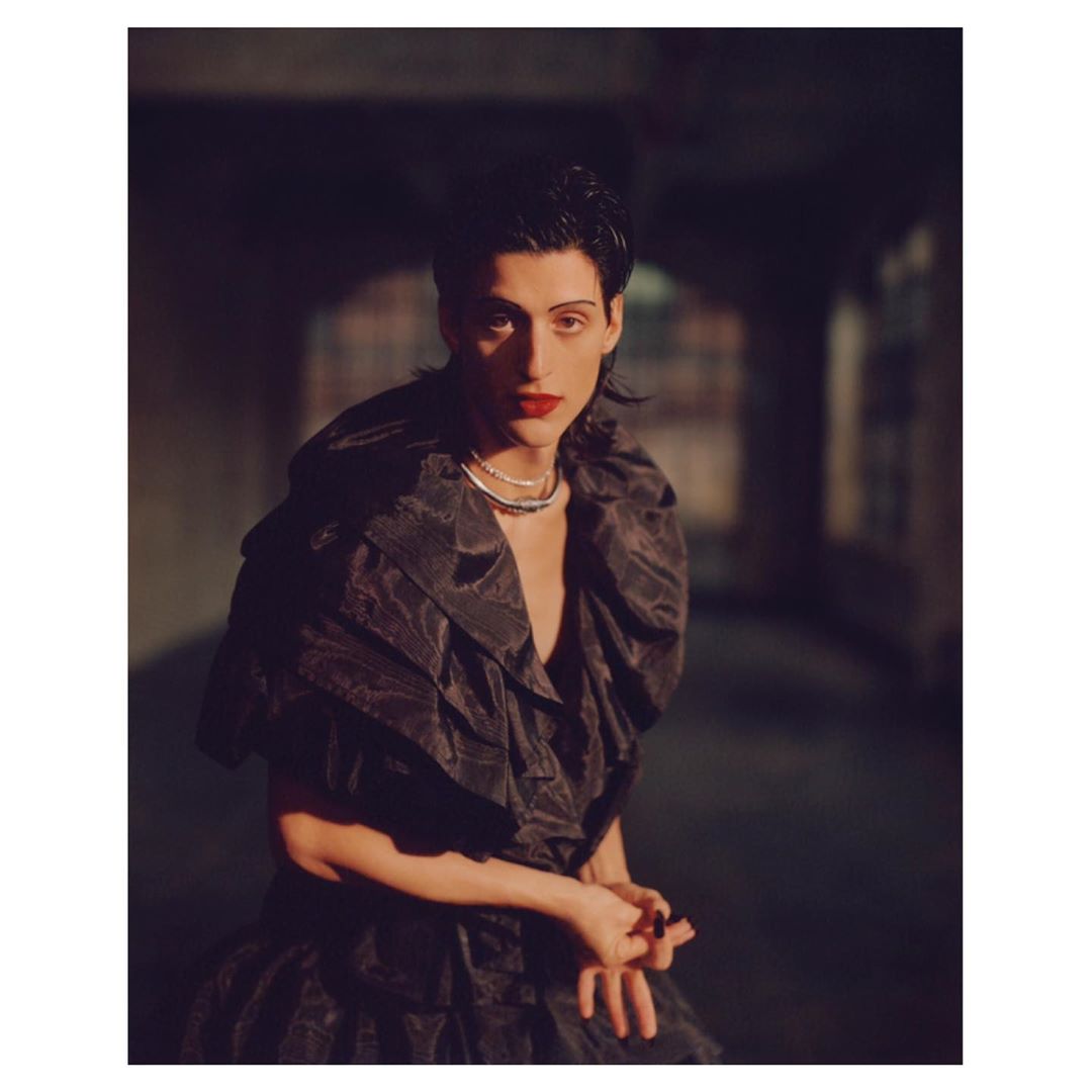 Portrait of a person with dark hair and red lipstick wearing a layered black ruffled dress in an arched hallway with dark lighting.