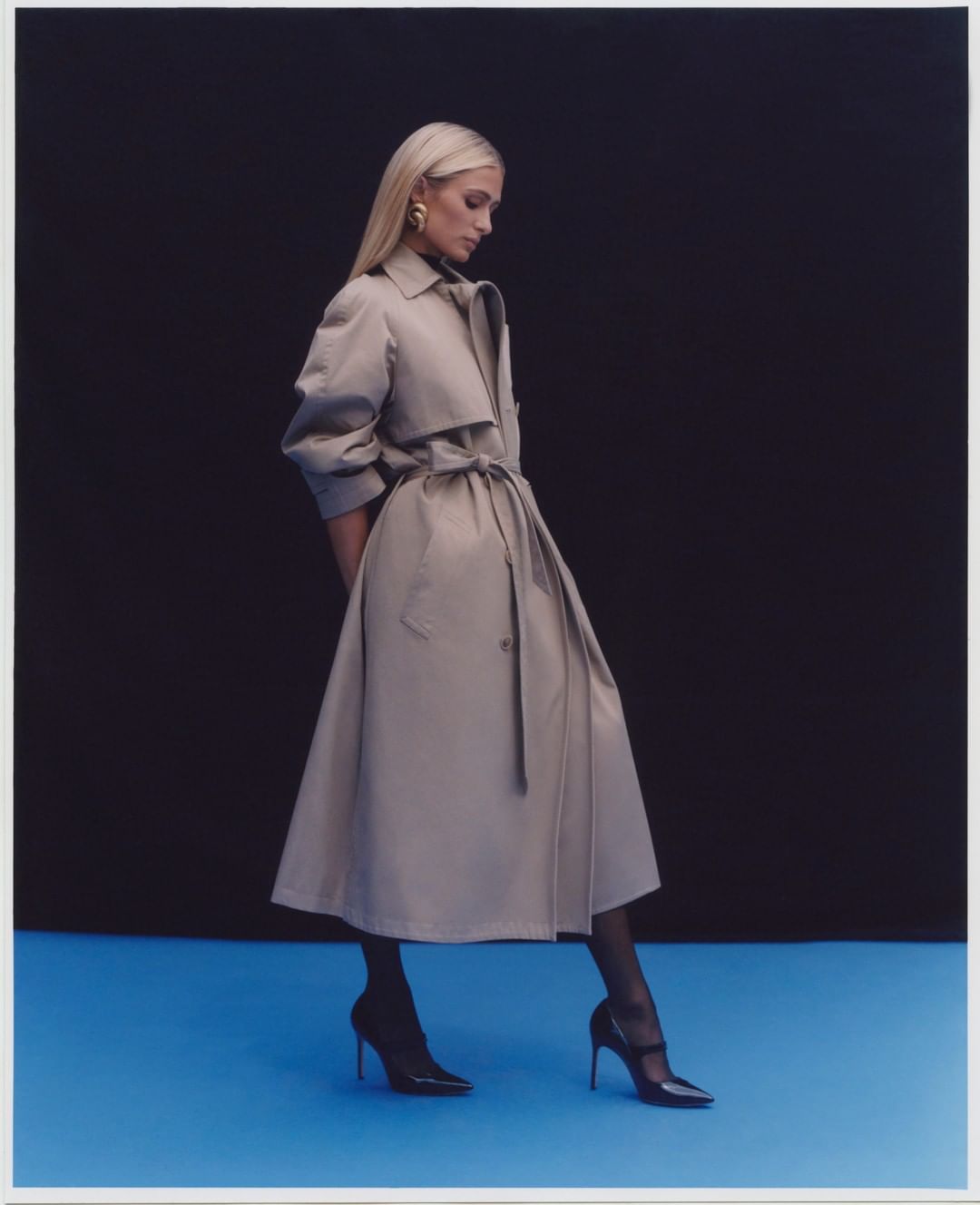Blonde woman wearing a tan trench coat and black heels standing against a dark backdrop over a blue floor.