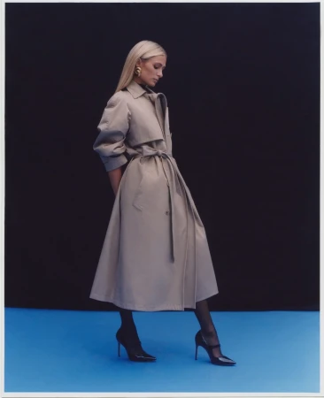 Blonde woman wearing a tan trench coat and black heels standing against a dark backdrop over a blue floor.