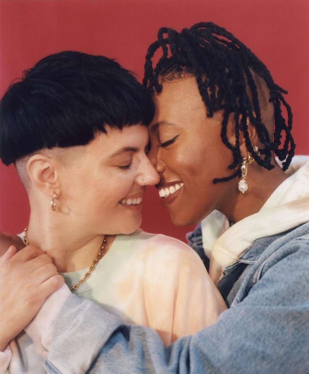 Two women embracing closely against a red backdrop. One woman has short black hair, while the other has dark skin and long dreadlocks.