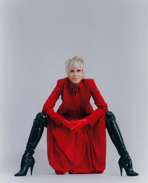 Adult woman in a red dress, blazer, and red gloves, posing with high-heeled, black, patent leather boots against a grey background.