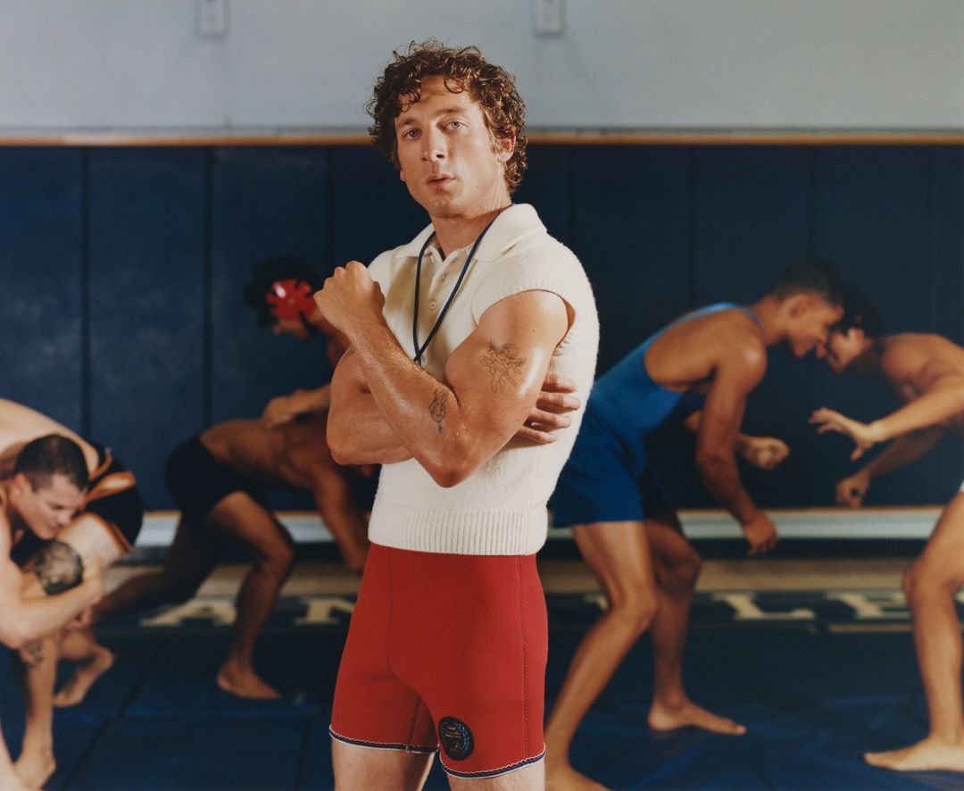 Man with arms crossed stands in a wrestling gym, with wrestlers in the background; he is wearing a polo shirt and red wrestling shorts.