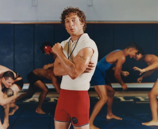 Man with arms crossed stands in a wrestling gym, with wrestlers in the background; he is wearing a polo shirt and red wrestling shorts.