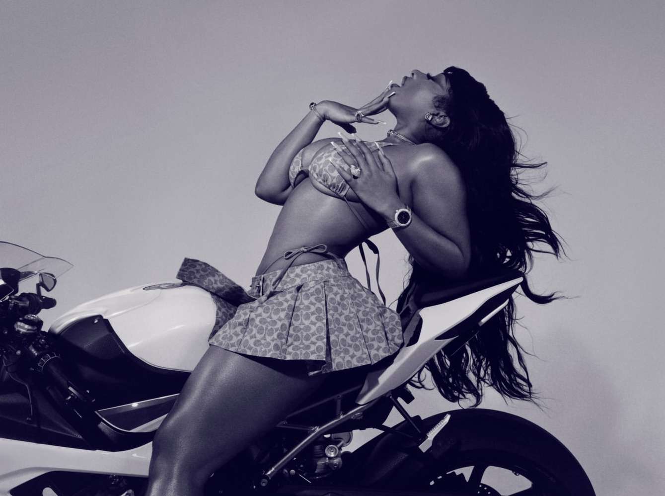 Woman in bikini and skirt sits on a motorcycle, posing in front of a solid light background in a glamour style photograph.