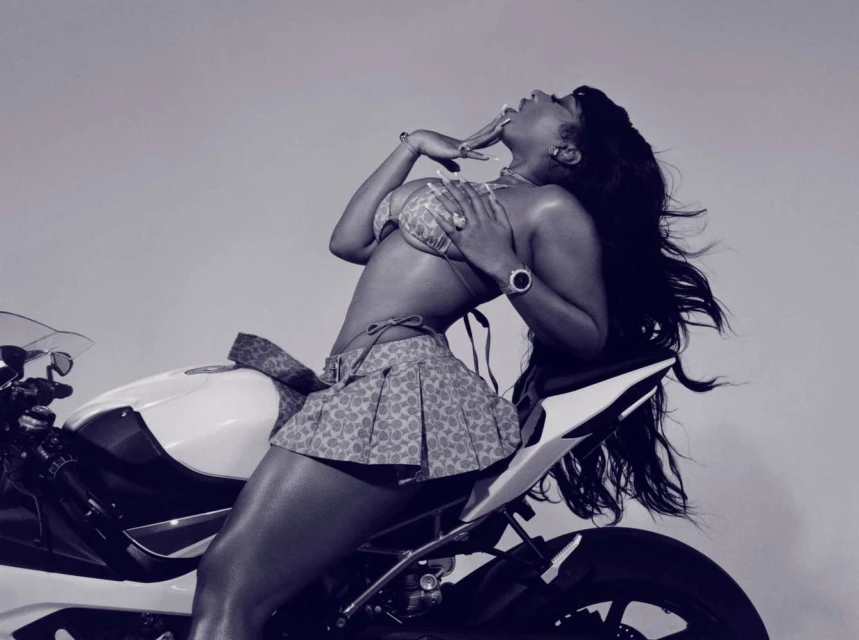Woman in bikini and skirt sits on a motorcycle, posing in front of a solid light background in a glamour style photograph.