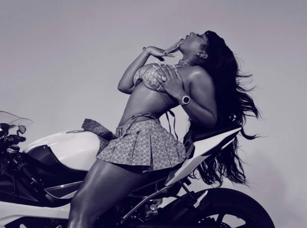 Woman in bikini and skirt sits on a motorcycle, posing in front of a solid light background in a glamour style photograph.
